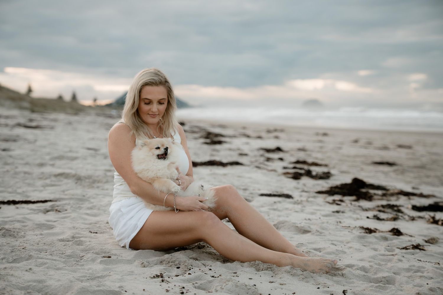 Blonde woman in white dress sitting on beach sand holding a small white fluffy dog at sunset.