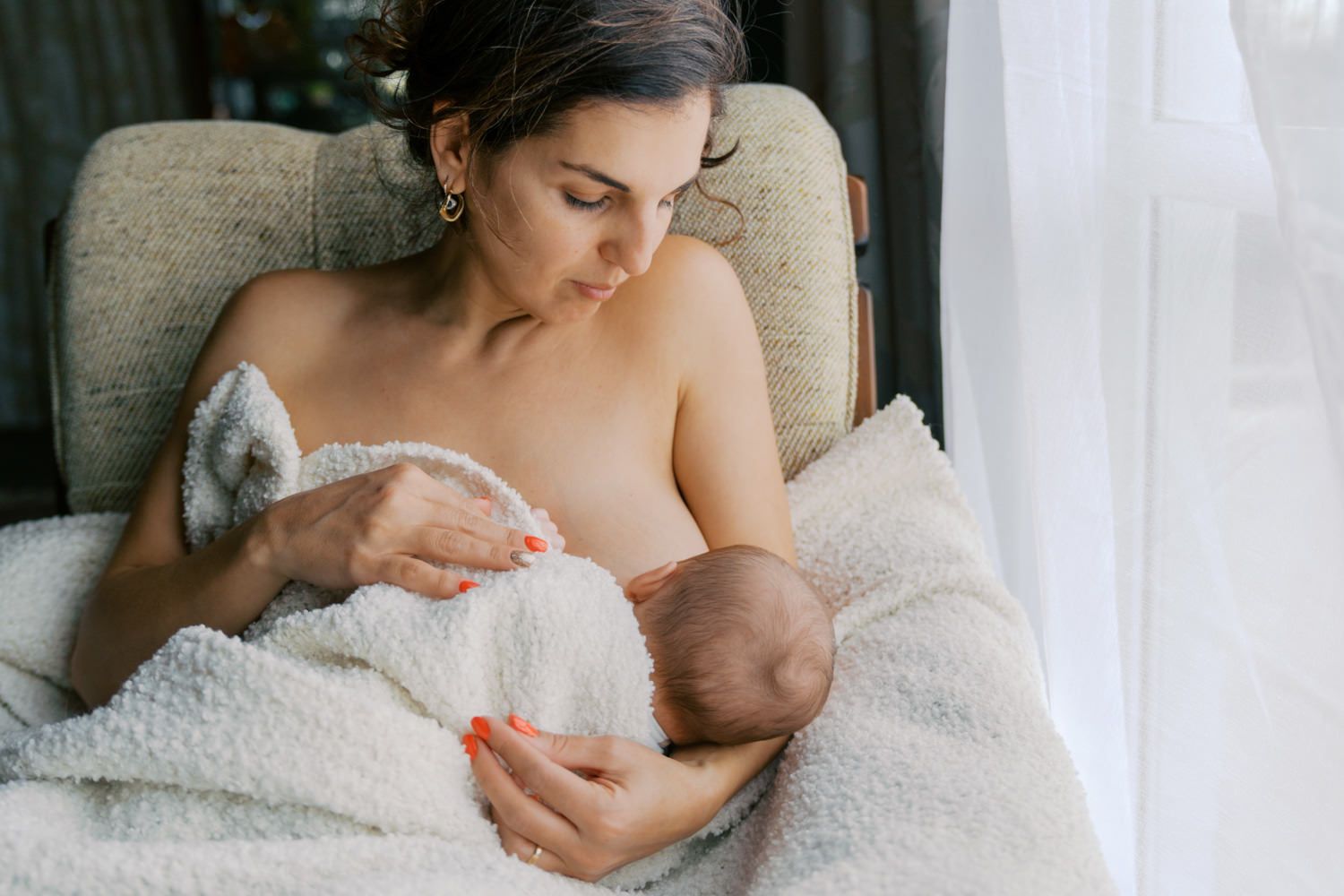 Mother breastfeeding her newborn baby while seated in a cozy chair covered with a white blanket.
