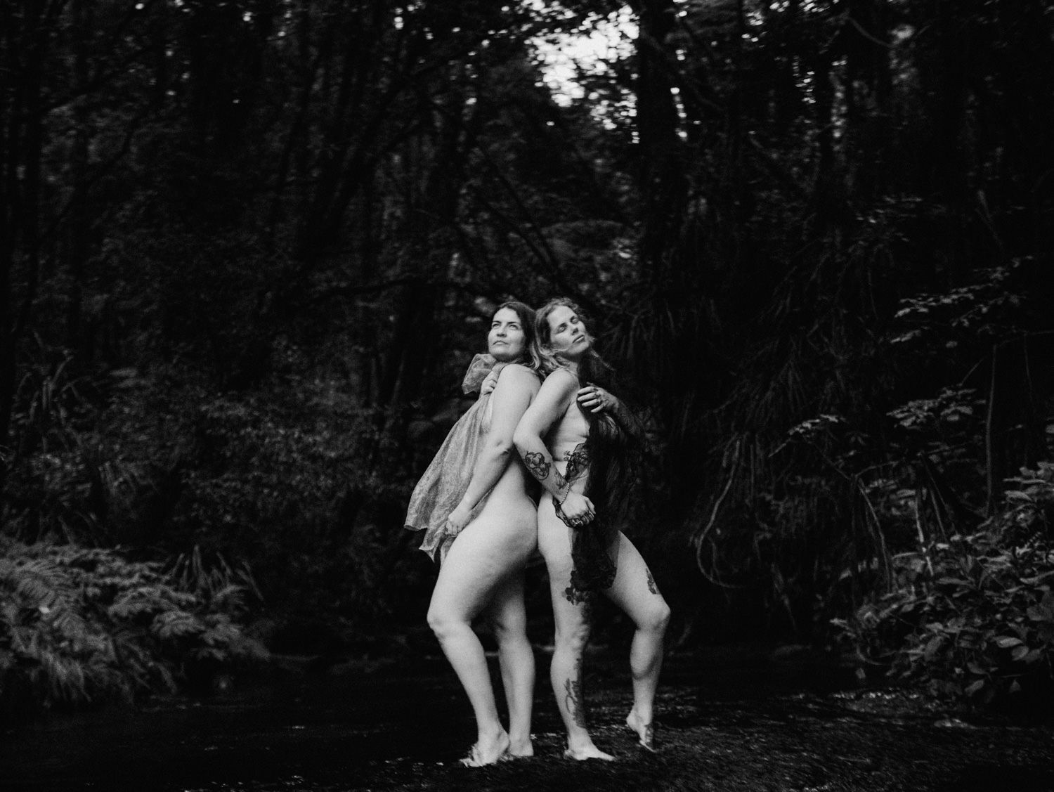 Two women pose back-to-back in a lush forest setting in a black and white fine art photograph.