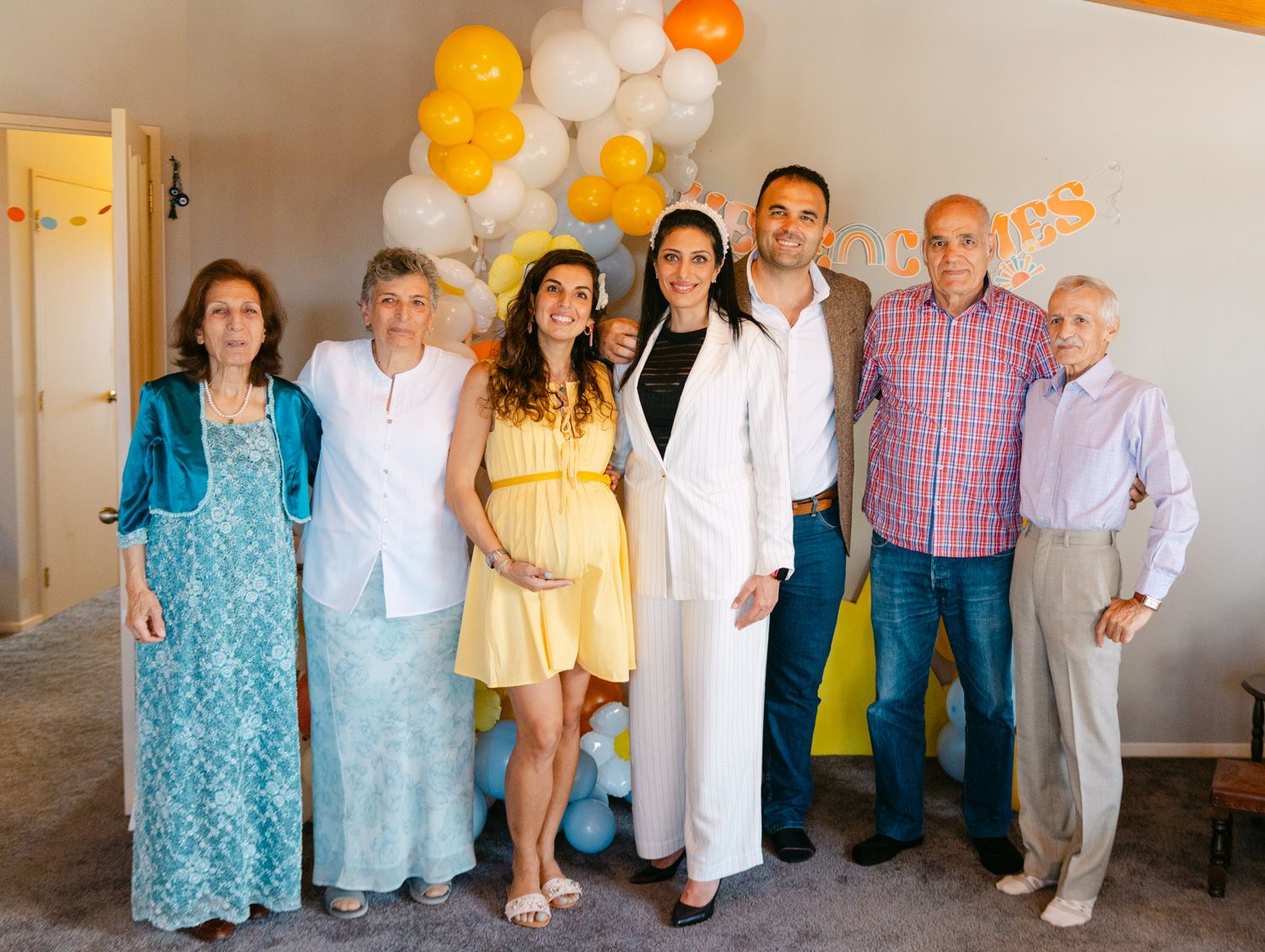 Family group photo at a baby shower with balloons, featuring a pregnant woman in yellow dress among loved ones.