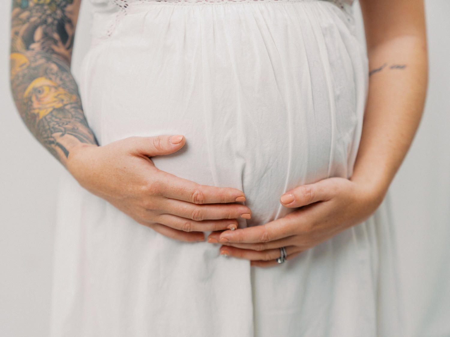 Pregnant woman in white dress cradling baby bump, with tattooed partner hands resting on belly.