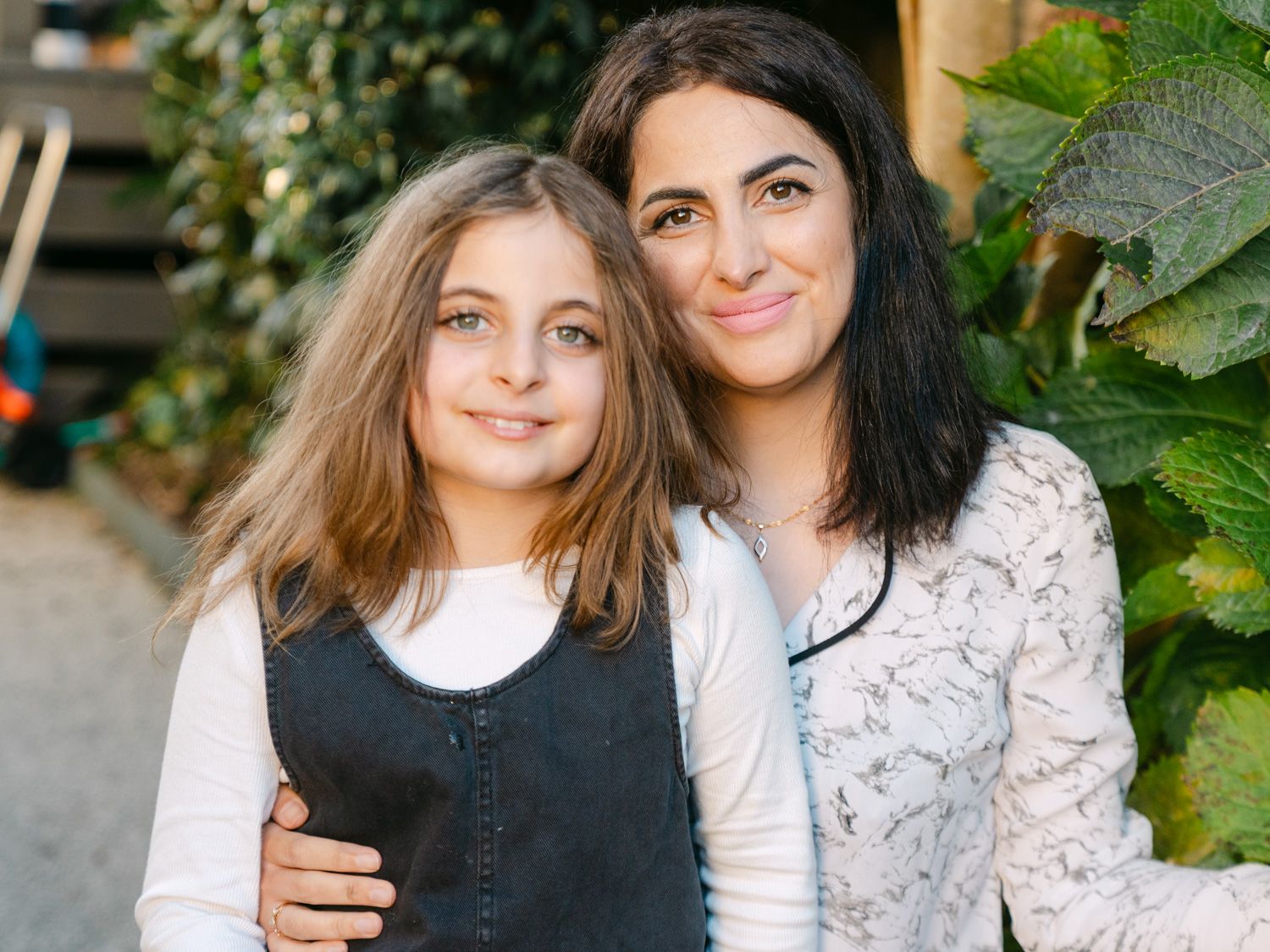 A woman and young girl smile together outdoors, posing for a portrait surrounded by green foliage.