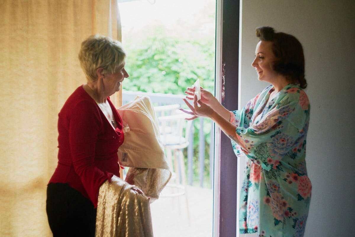 Two women chat near a bright doorway, one in red holding a shawl, the other in a floral robe holding a gift.
