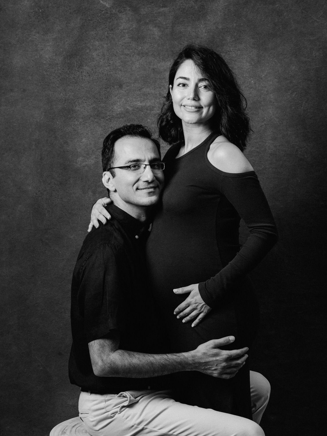 Black and white maternity photo of a couple, man seated with hand on pregnant womans belly, studio portrait.