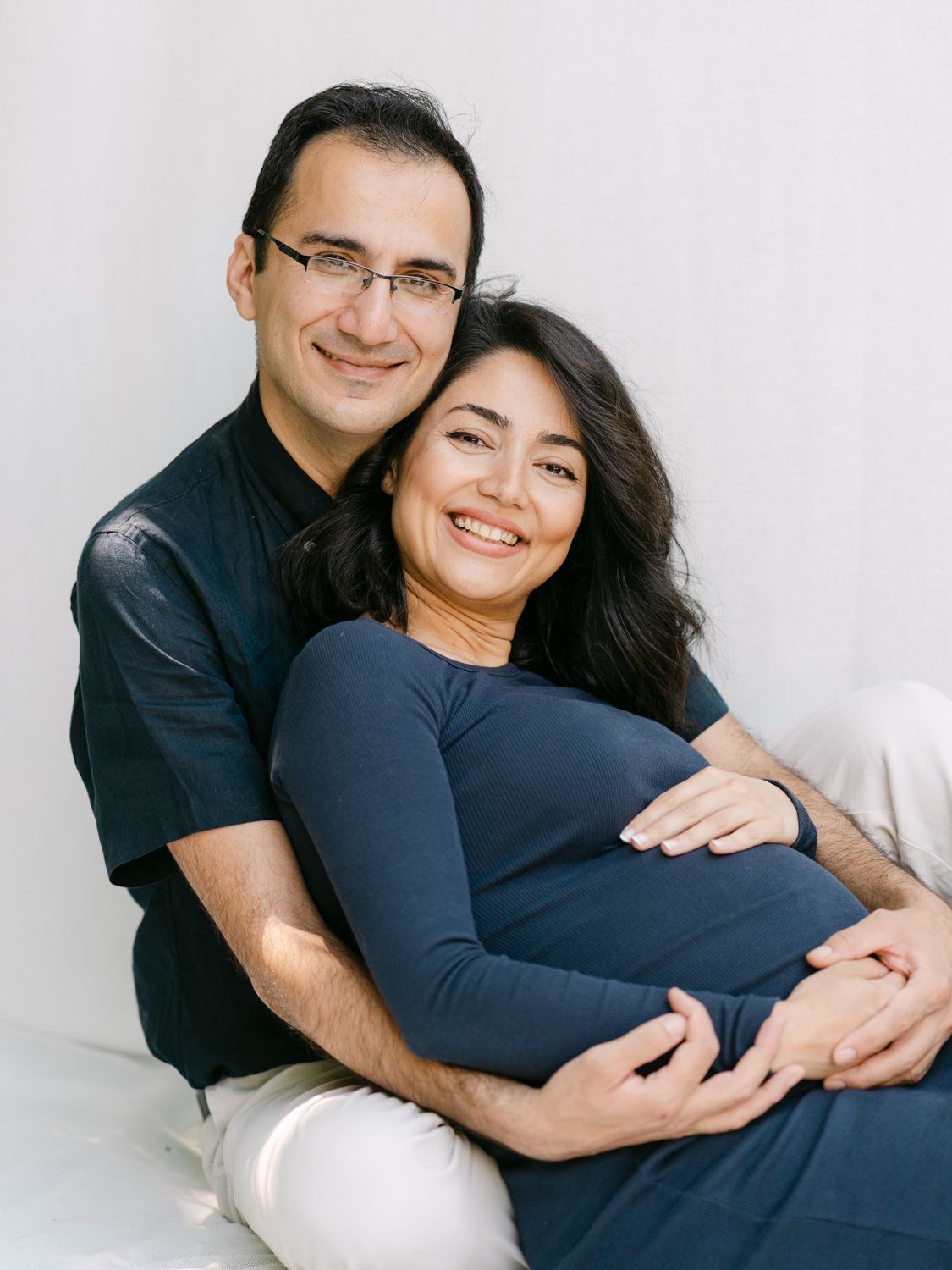 Smiling couple posing for maternity photo, man embracing pregnant woman in navy dress against white background.