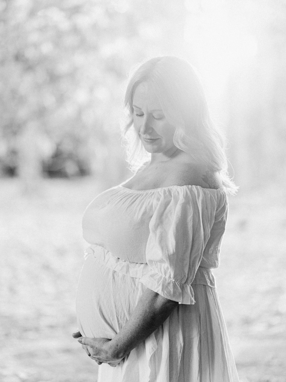 Black and white maternity photo of a woman in an off-shoulder dress cradling her baby bump outdoors.