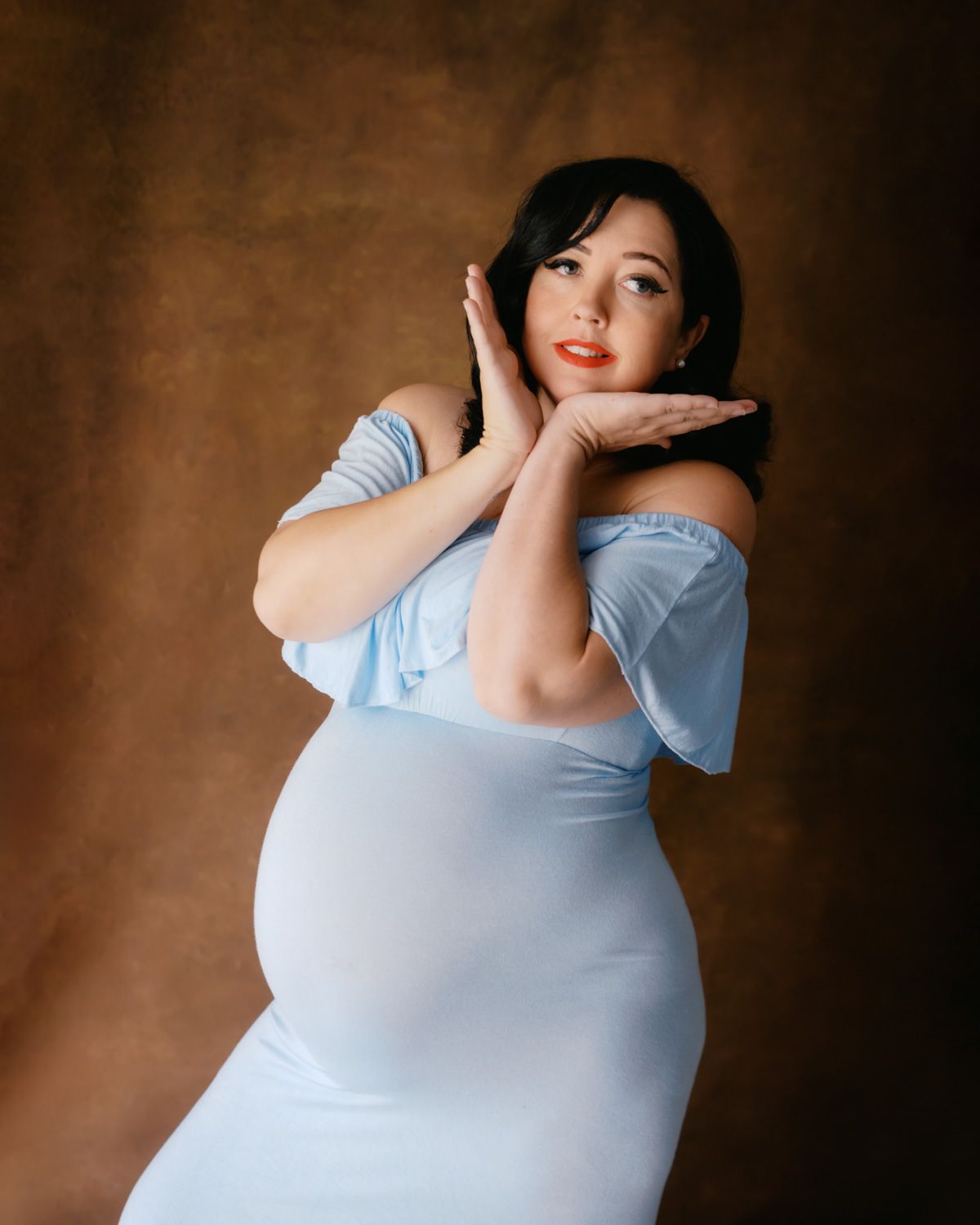 Pregnant woman in light blue off-shoulder dress posing gracefully with hands near face against brown backdrop.