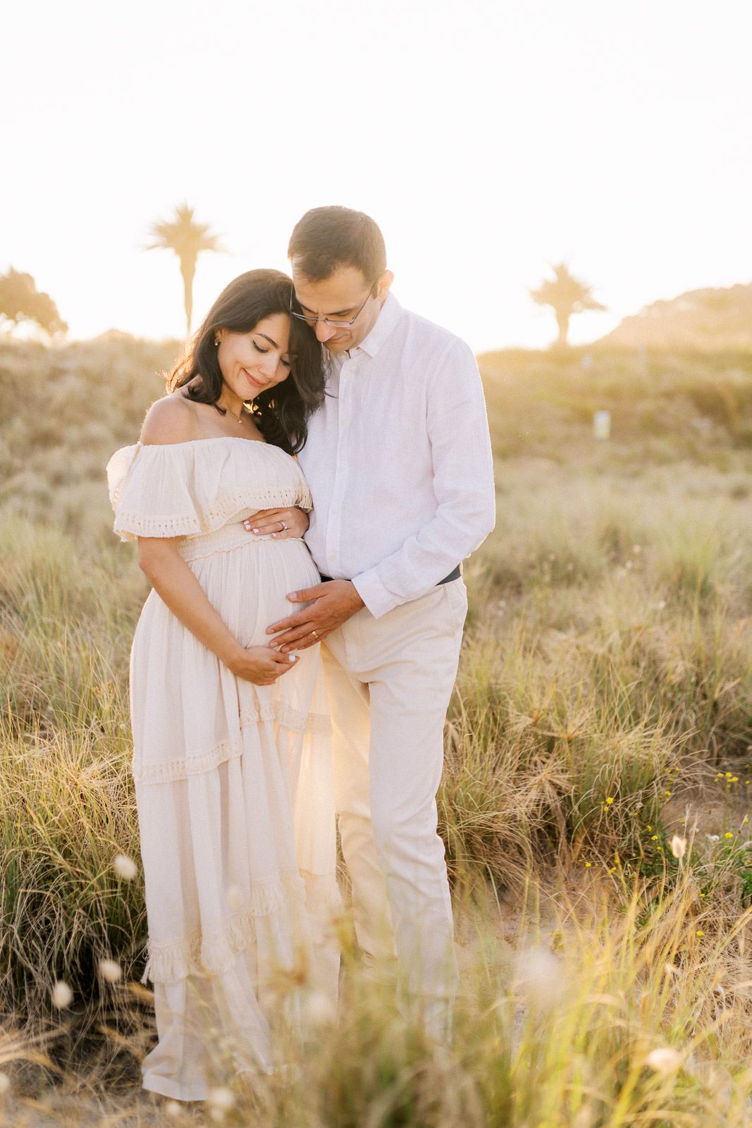 Couple tenderly cradling pregnant belly at golden hour in a sunlit field with palm trees in background.
