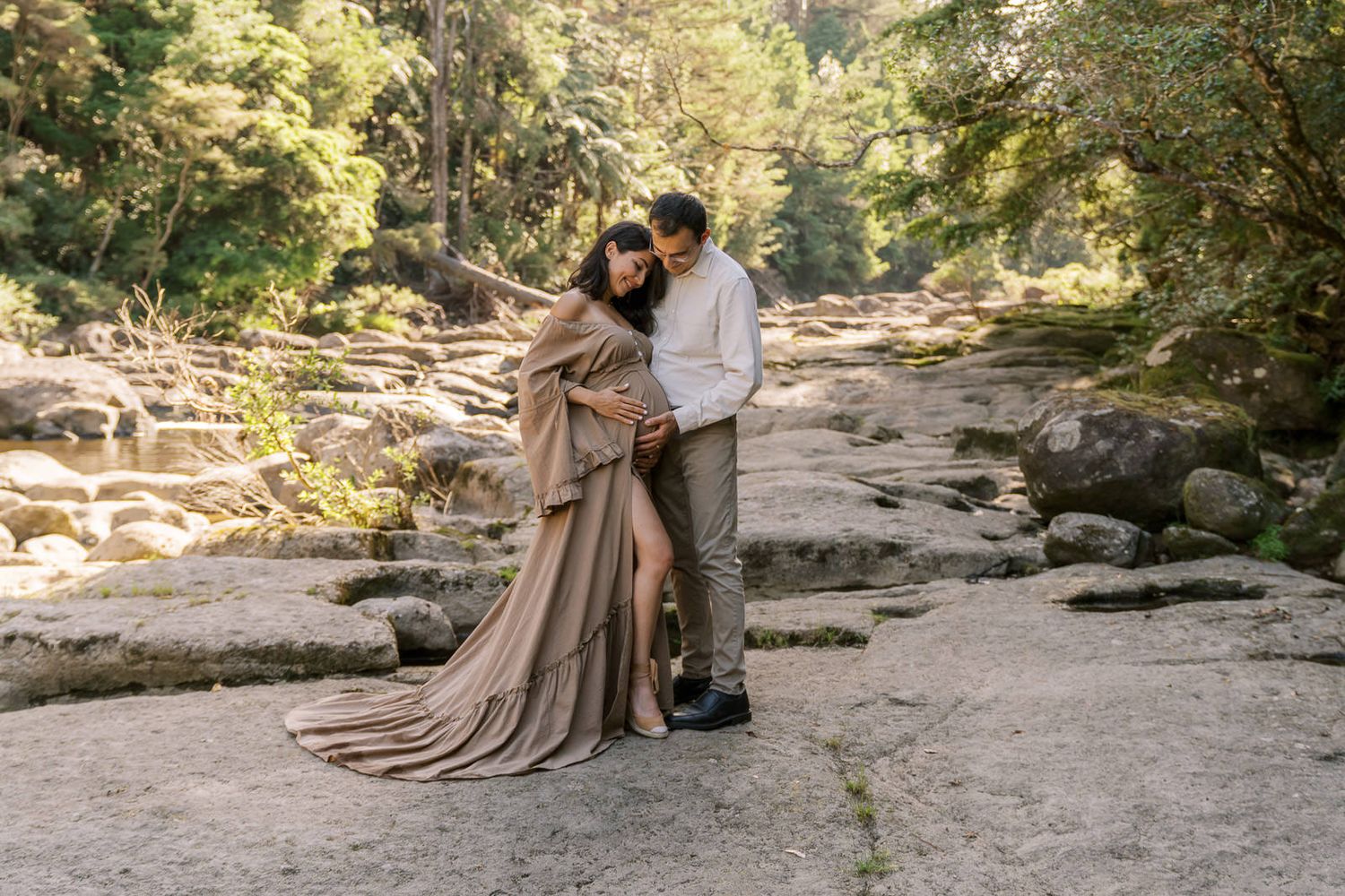 Couple embracing on rocky riverbed in forest, woman in flowing tan maternity gown, man in white shirt.