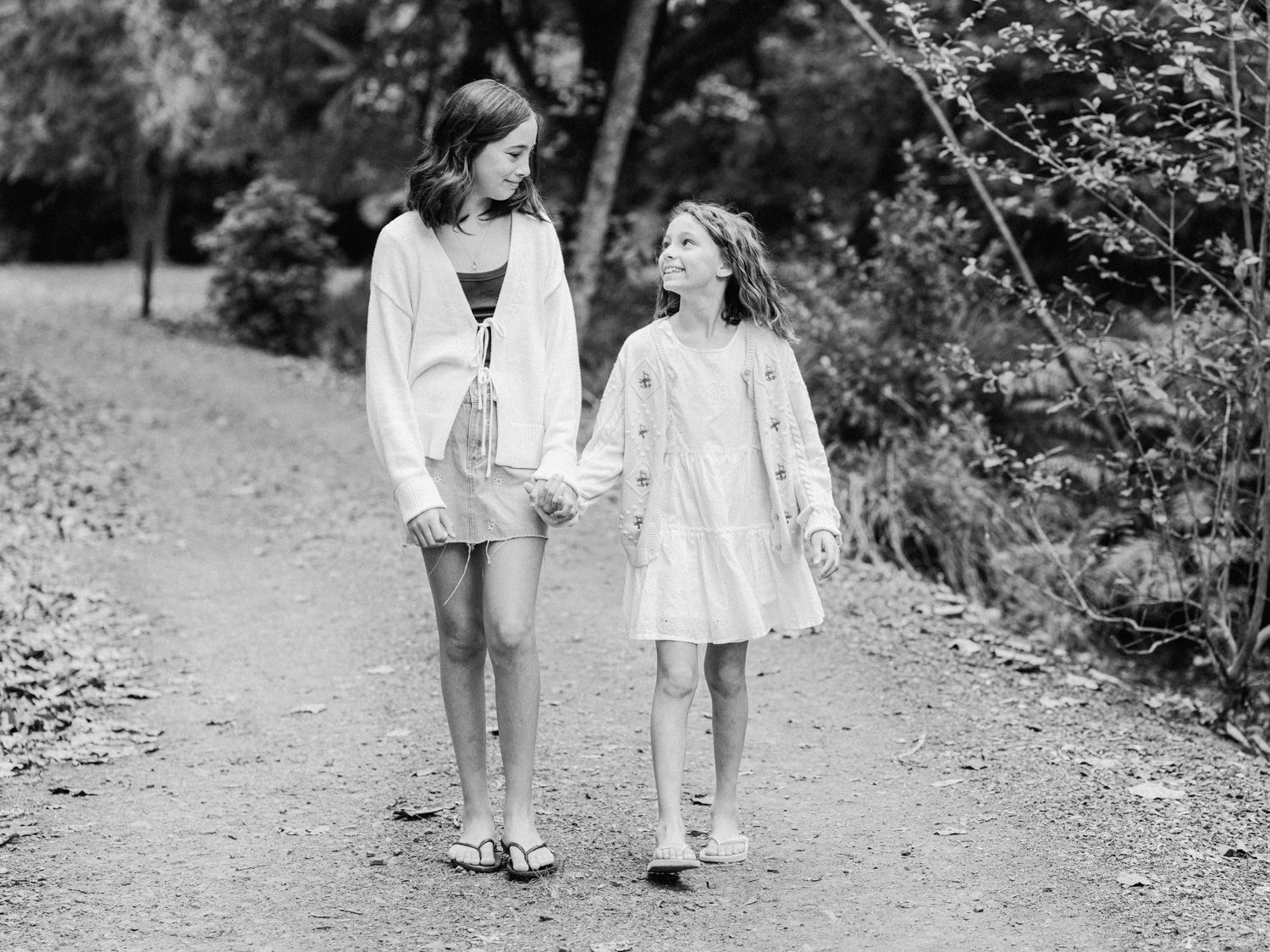 Two girls holding hands and smiling while walking on a tree-lined path, black and white photo.