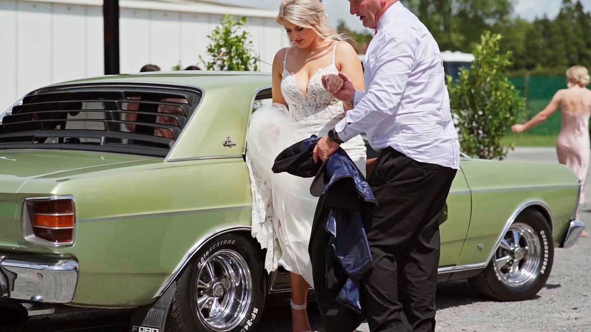 Bride in white lace gown assisted by man in suit near vintage green muscle car at outdoor wedding.