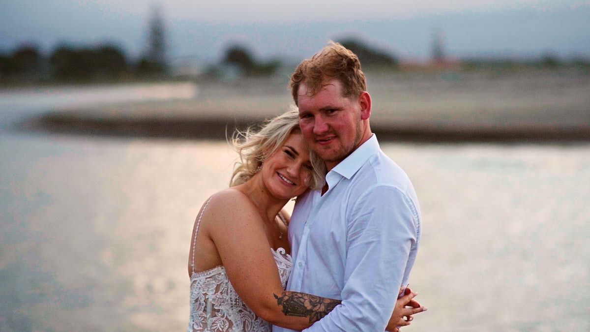 Couple embracing romantically at sunset by a calm waterfront, woman in floral dress, man in light blue shirt.