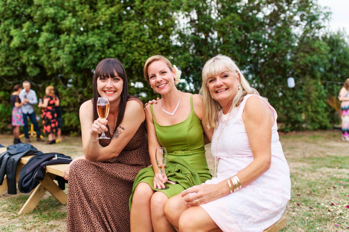 Three women smiling and posing together outdoors at a garden party, one holding a champagne glass.