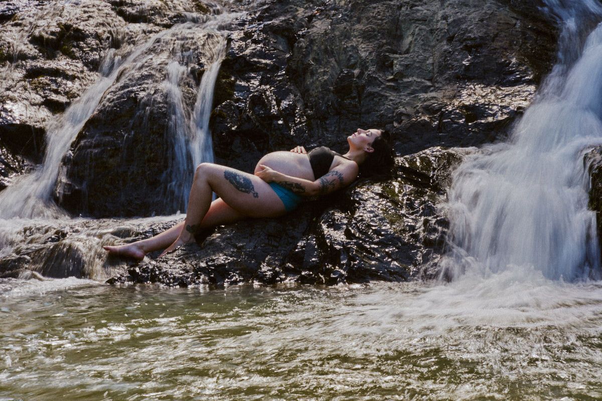 Woman in blue bikini relaxing on rocks beside a cascading waterfall in a tropical outdoor setting.