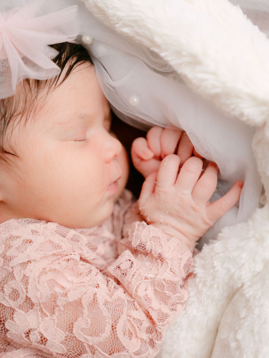 Sleeping newborn baby in pink lace outfit with bow headband, nestled in soft white blanket.
