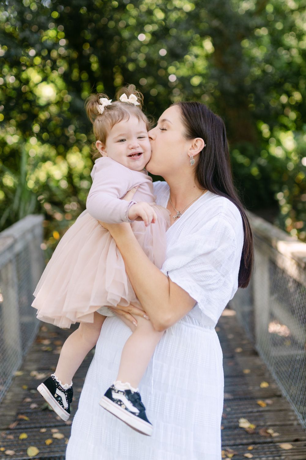 A mother in a white dress kisses her smiling toddler daughter in a pink tutu dress outdoors on a bridge.