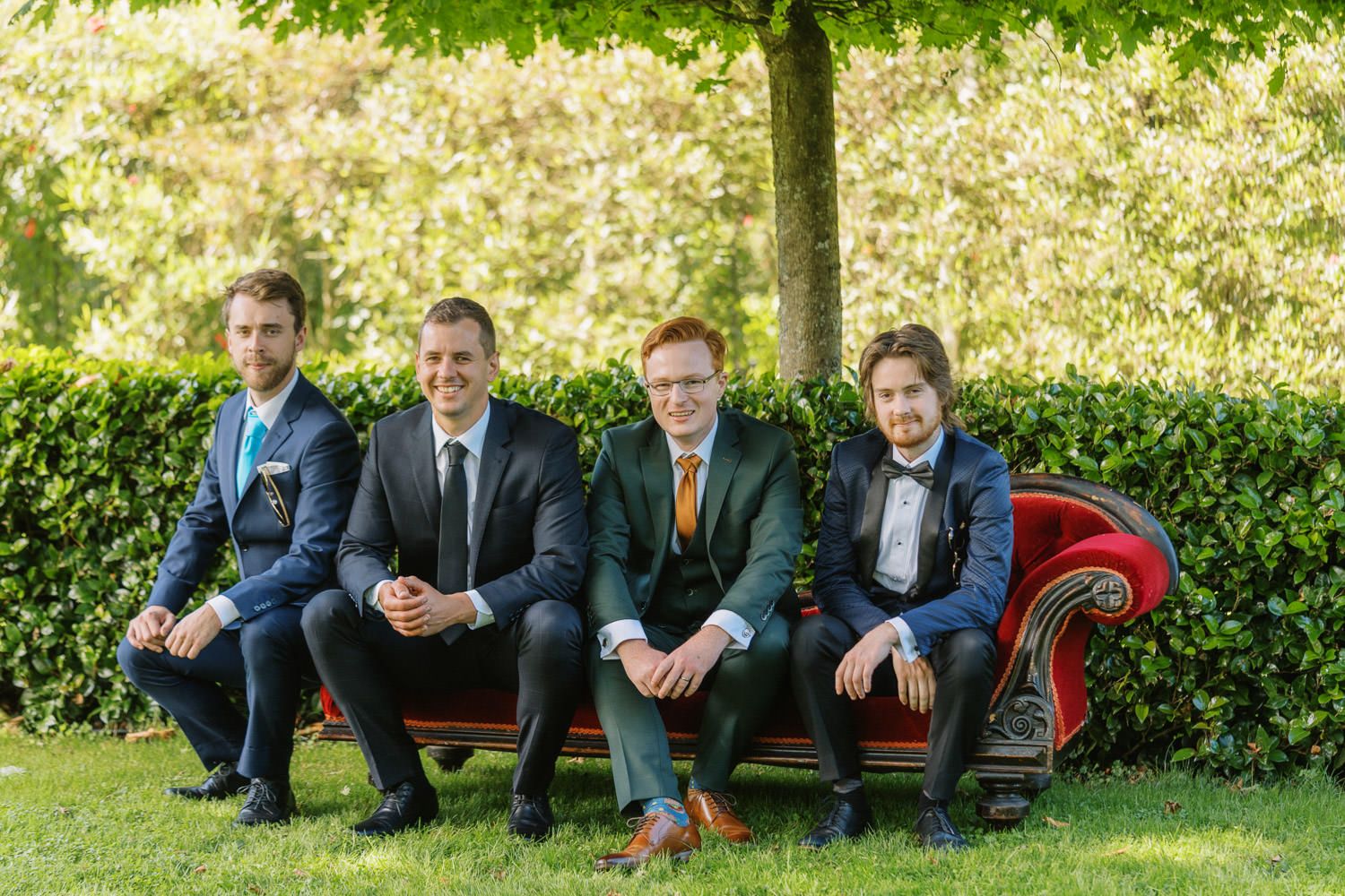 Four men in formal suits sitting on a red vintage bench outdoors under a tree on green grass.