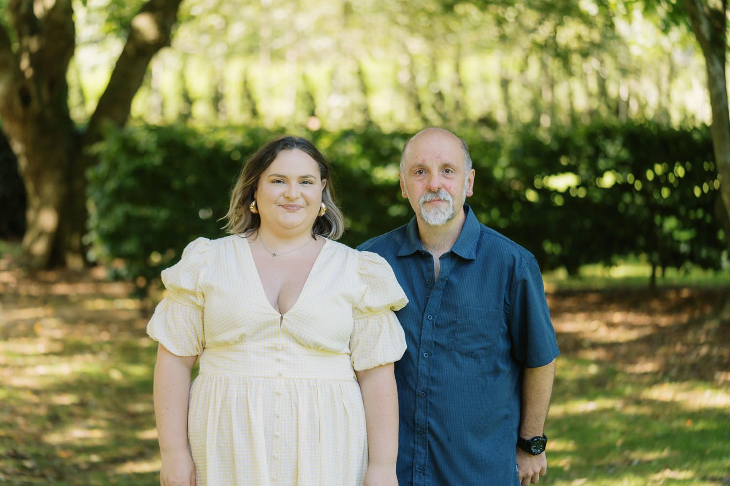 A smiling couple poses outdoors in a lush green park, woman in white dress and man in blue shirt.