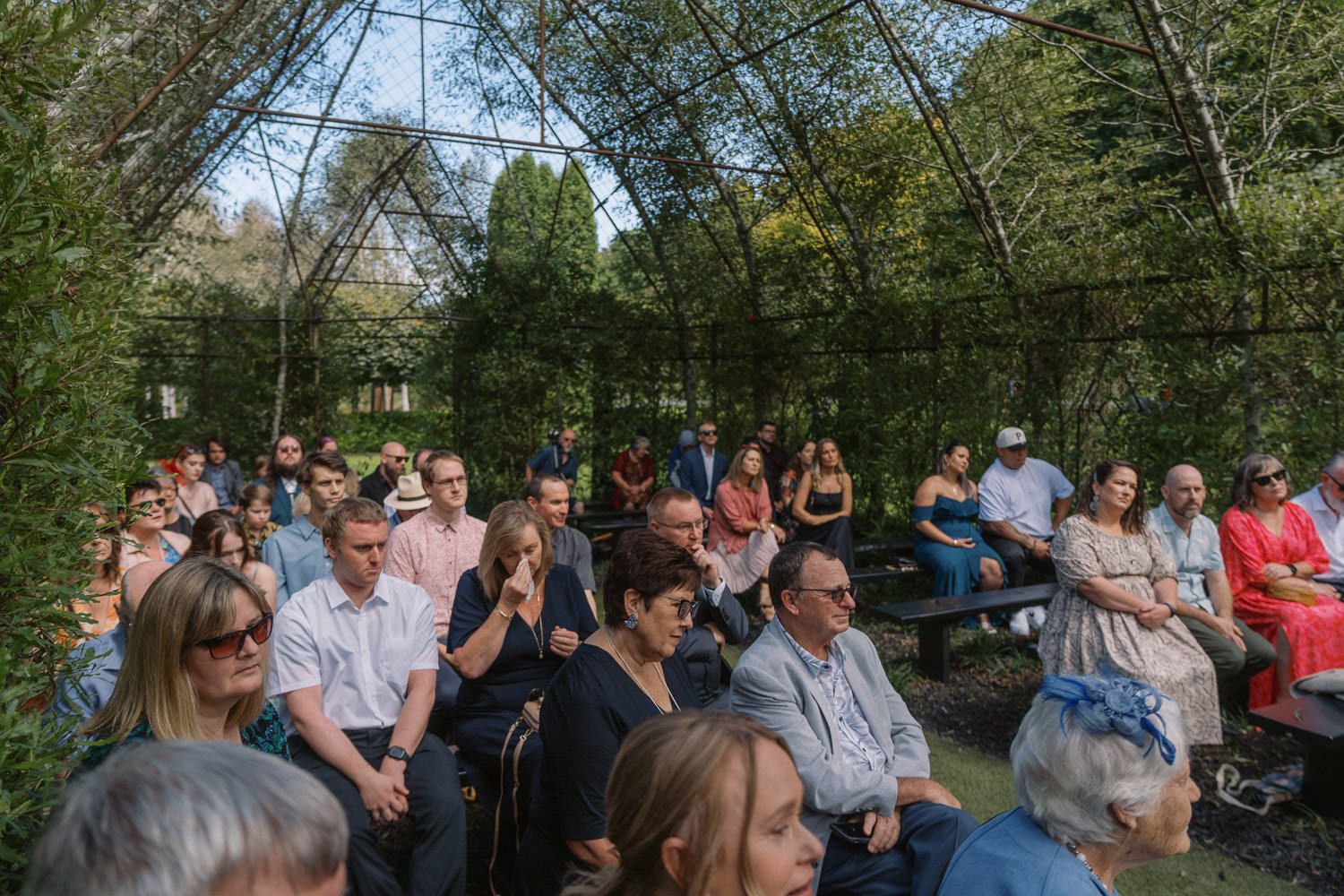Crowd of guests seated outdoors under a leafy green pergola structure at a summer garden event.