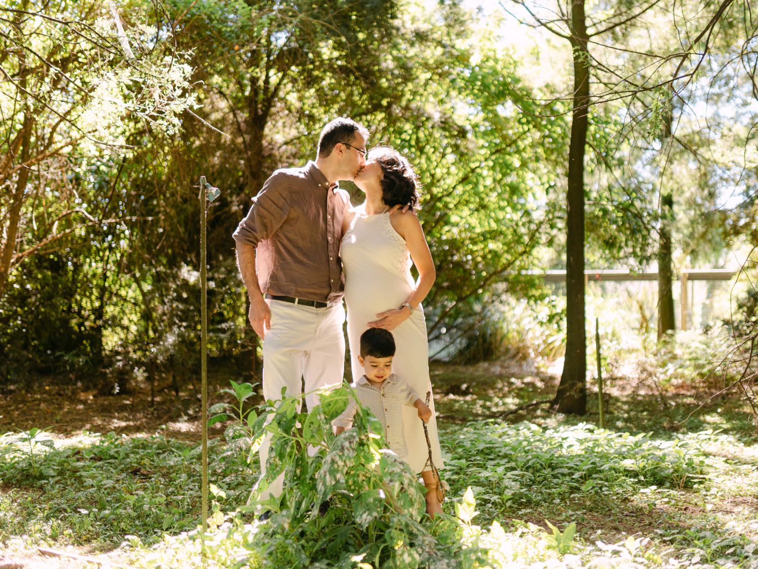 Pregnant couple kissing outdoors with young child standing between them in a lush green garden setting.
