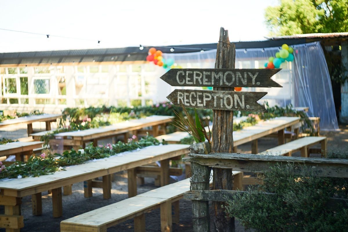 Rustic wooden directional signs pointing to ceremony and reception areas with farm tables and colorful balloon decorations.