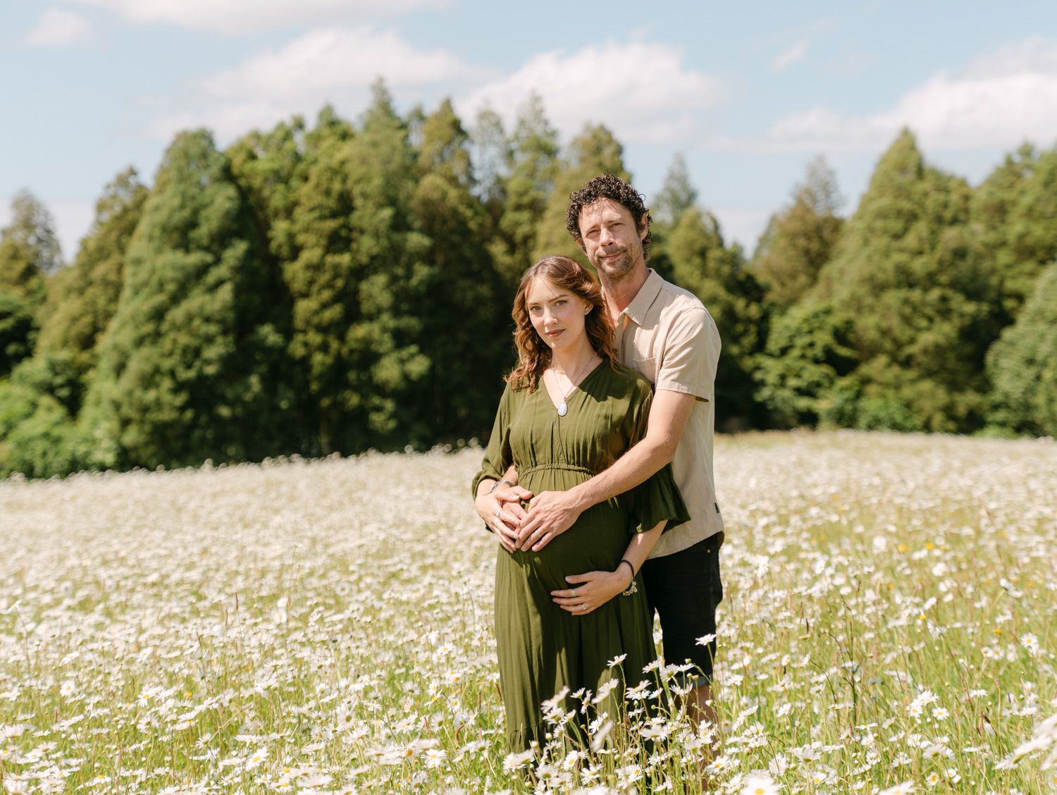 Pregnant couple standing in a white daisy field with green forest backdrop during a maternity photo shoot.