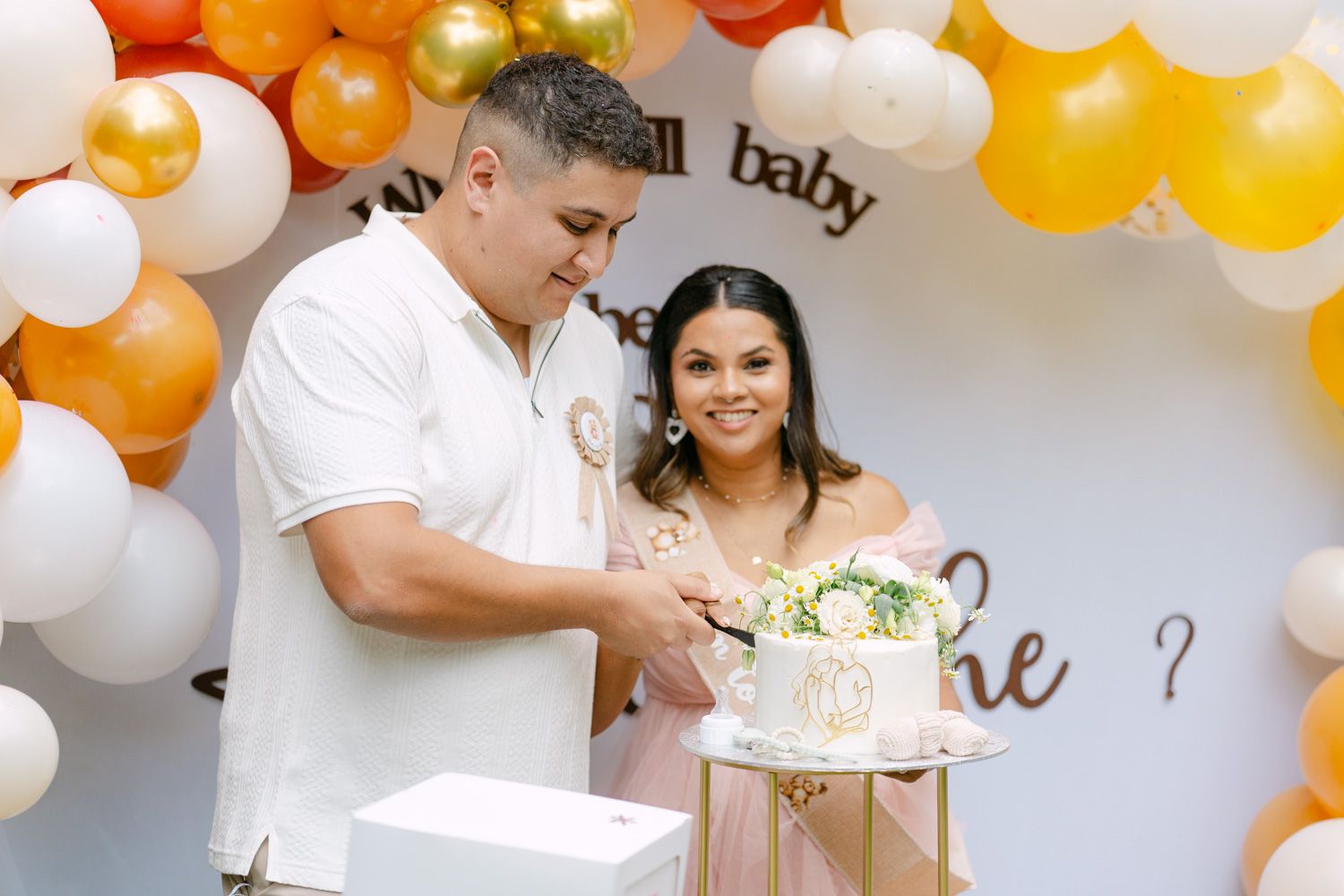Couple cutting cake at a gender reveal party with orange and white balloon arch backdrop.