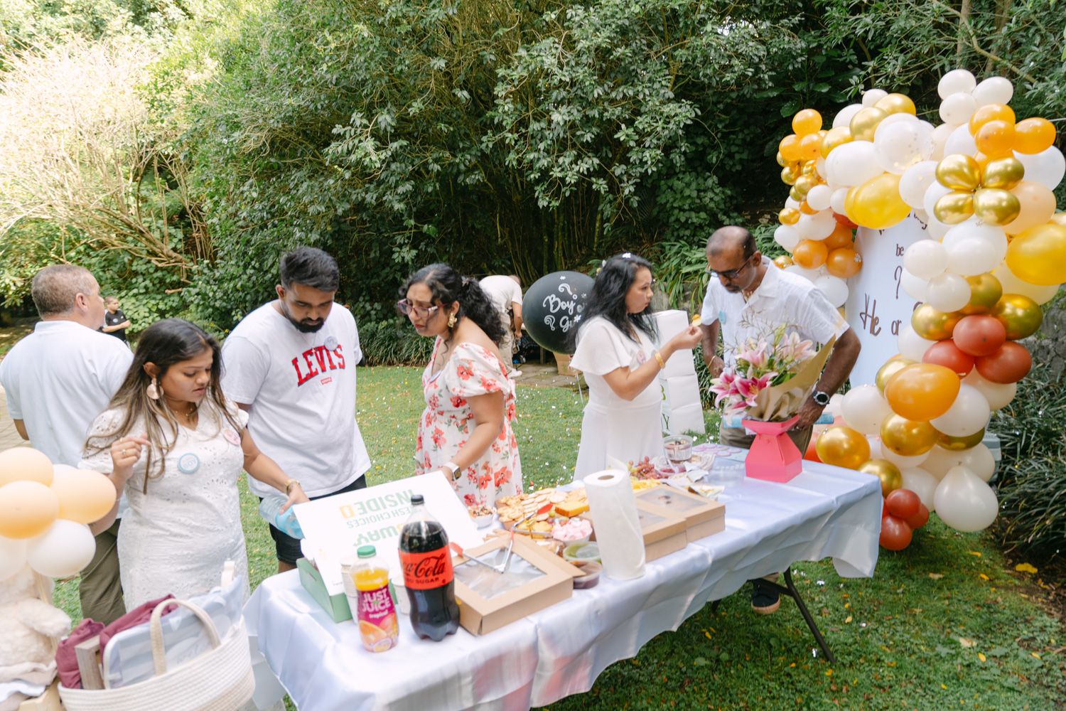 Outdoor birthday party with guests gathered around a decorated table with balloons, cake, and Coca-Cola in a lush garden.