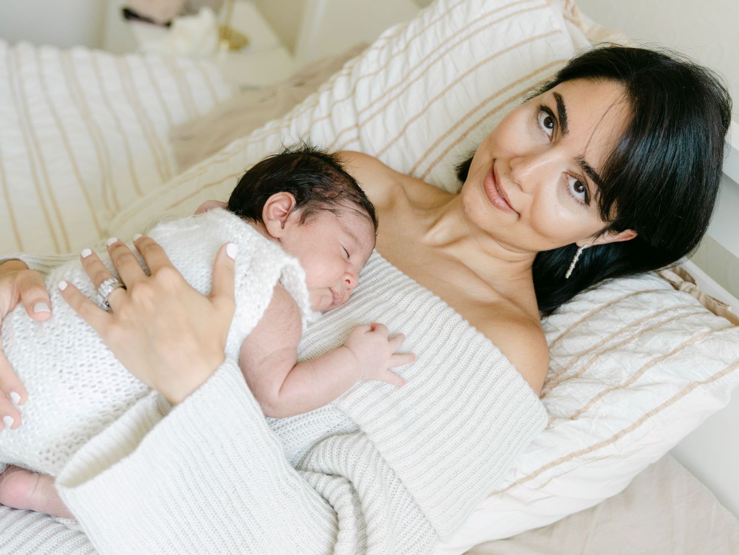 Smiling dark-haired mother lying on bed holding sleeping newborn baby in white outfit.