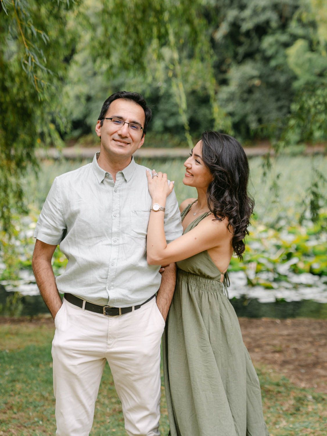 Couple posing outdoors near a pond with lily pads, woman in green dress resting hand on mans shoulder.