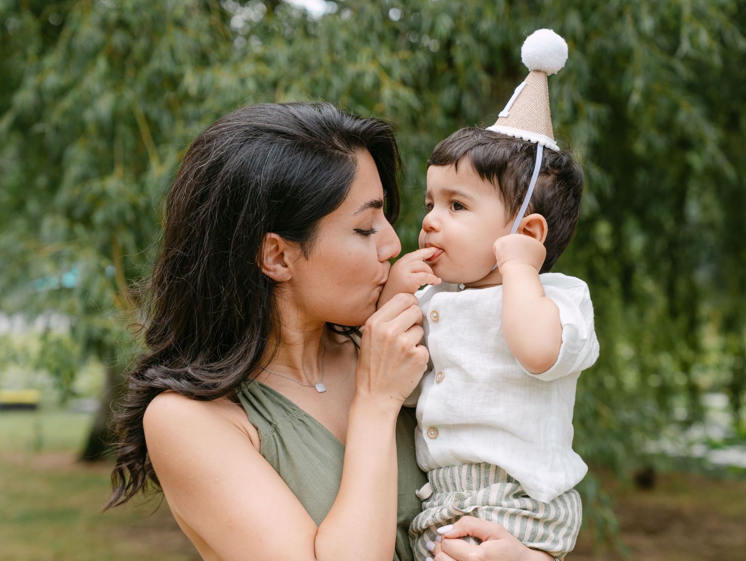 A mother kisses her baby wearing a party hat outdoors near green weeping willow trees.