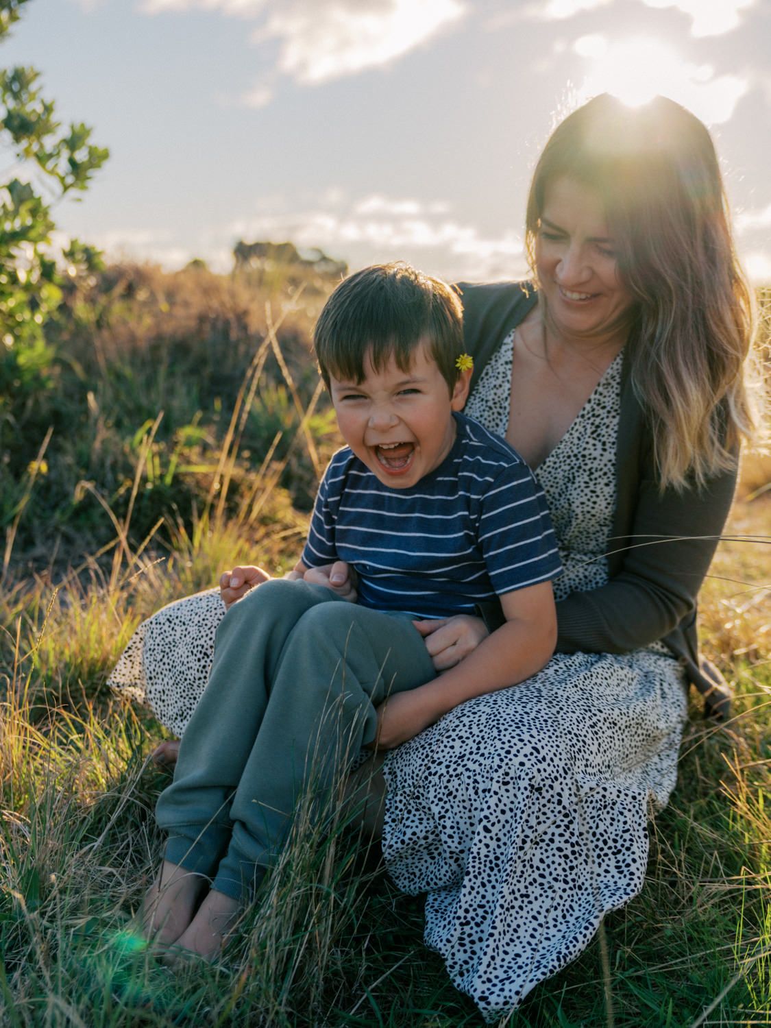 A joyful boy laughs while sitting on his smiling mother's lap in a sunlit grassy field at golden hour.