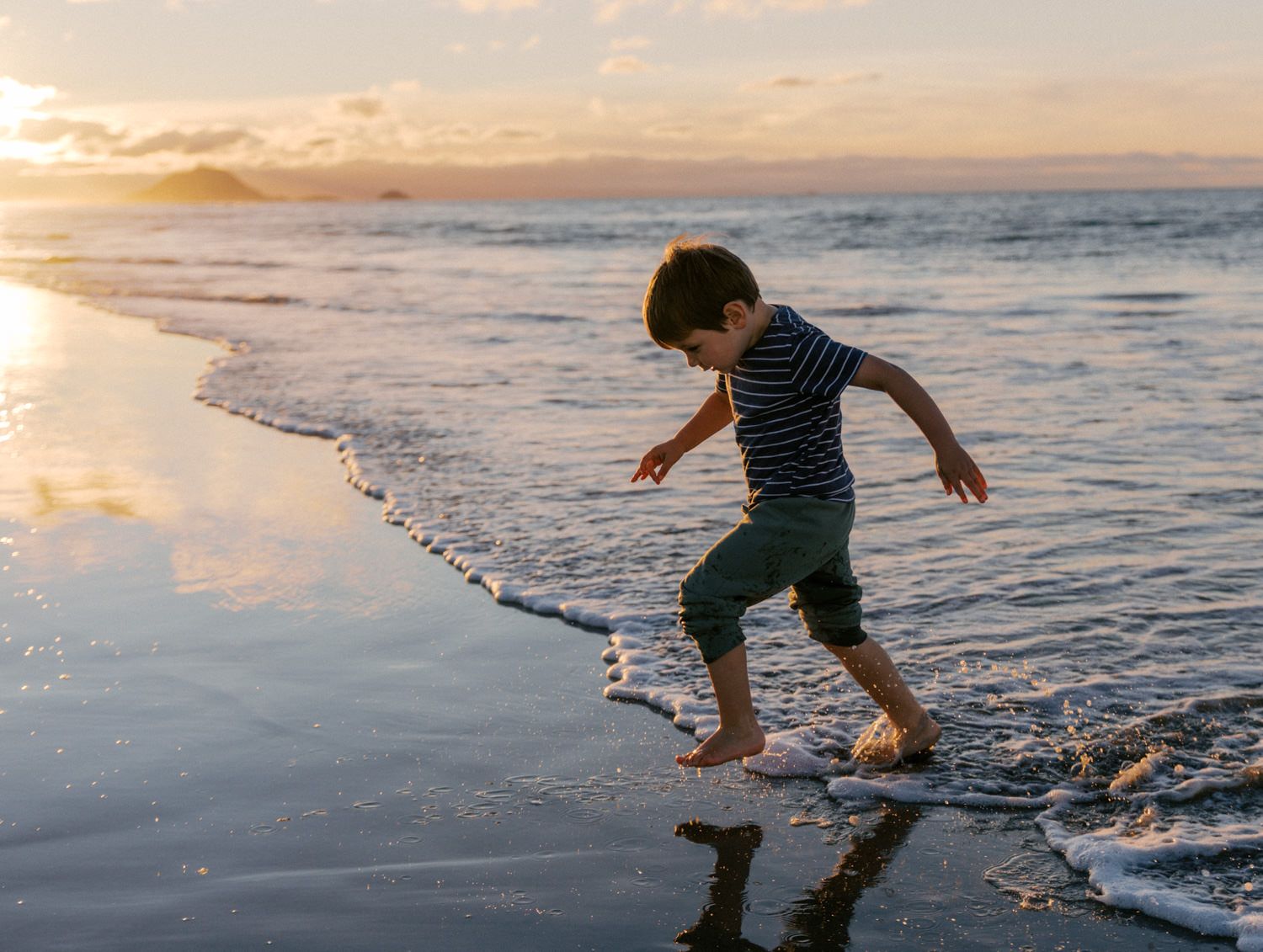 A young boy joyfully runs through gentle ocean waves at a beautiful golden sunset beach.