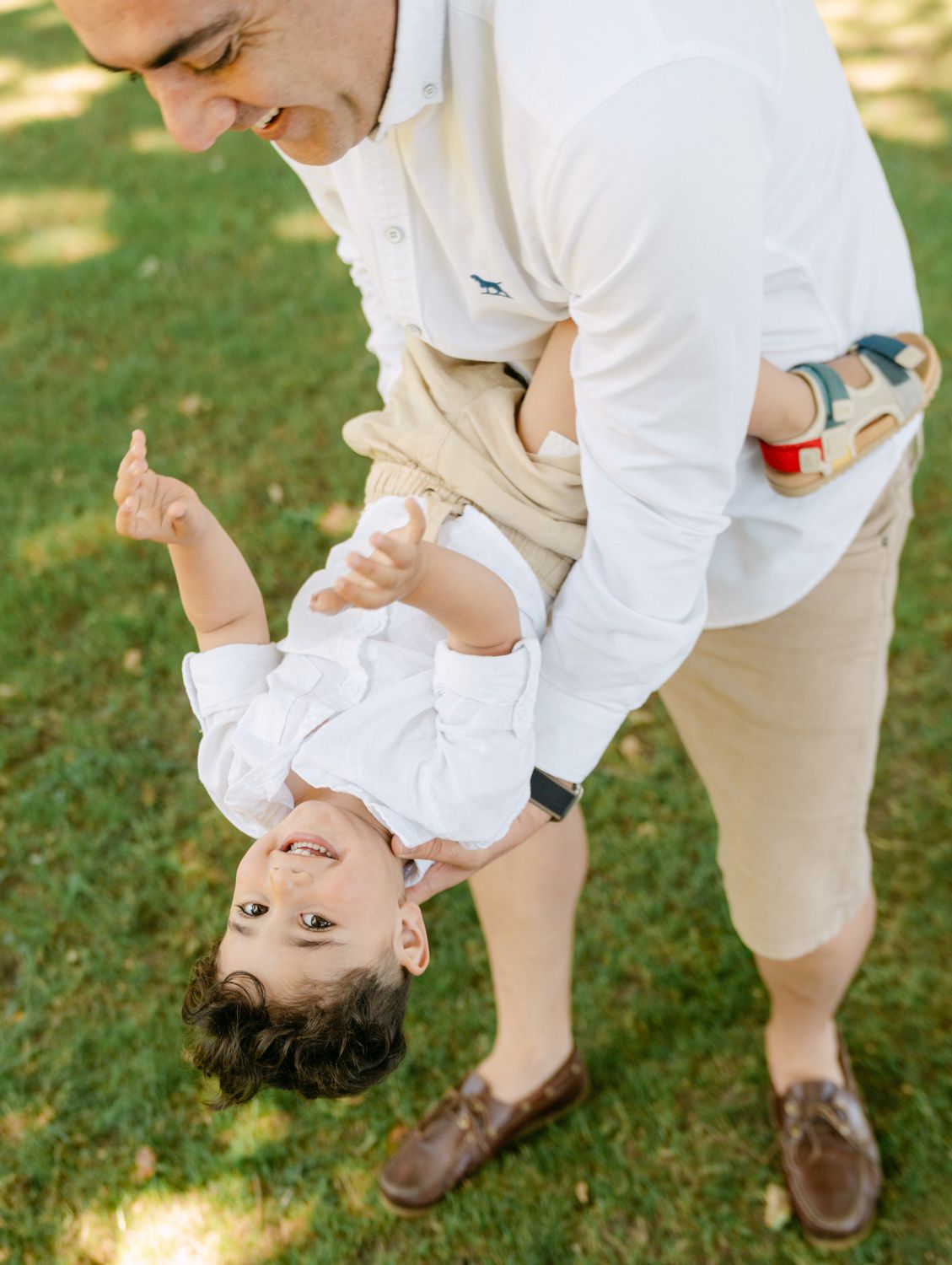 A father playfully holds his laughing toddler upside down on a green lawn, both dressed in white outfits.