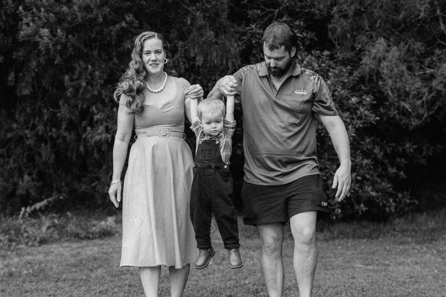 Black and white family photo of parents swinging young child between them on grass with trees in background.