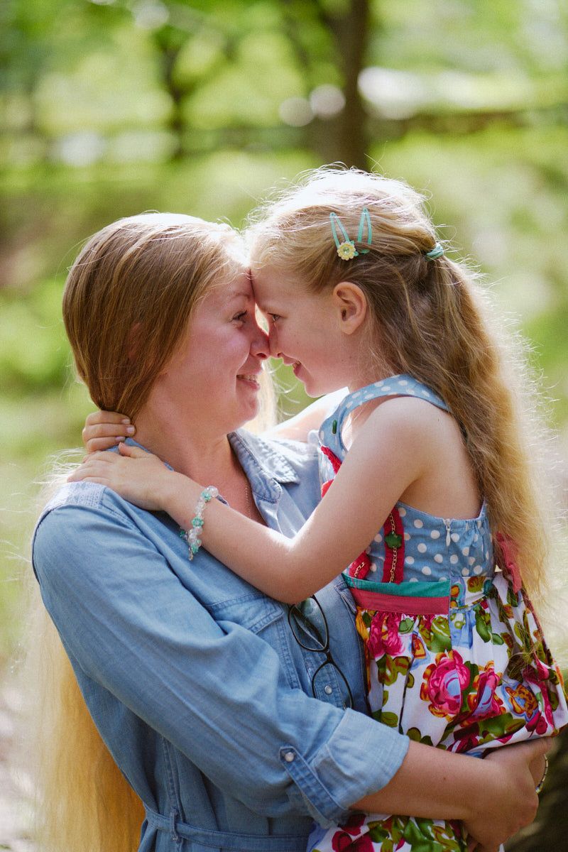 A mother and young daughter touch foreheads and smile while embracing outdoors in a lush green park setting.