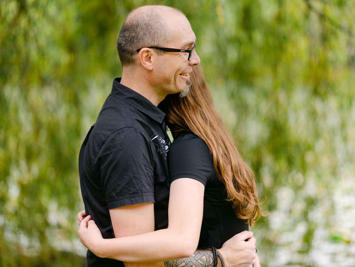 A bald man with glasses embraces a woman with long brown hair outdoors near a willow tree and pond.