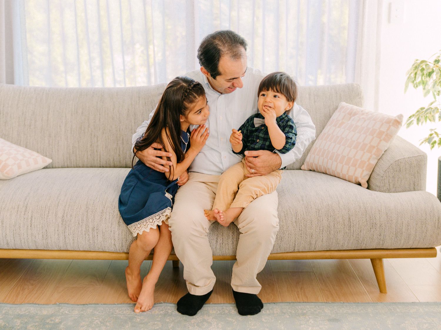 A father sits on a gray sofa with his two young children, sharing a warm family moment at home.