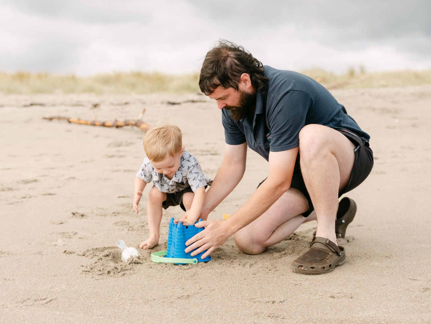 Father and toddler playing with blue bucket in sand on overcast beach day.