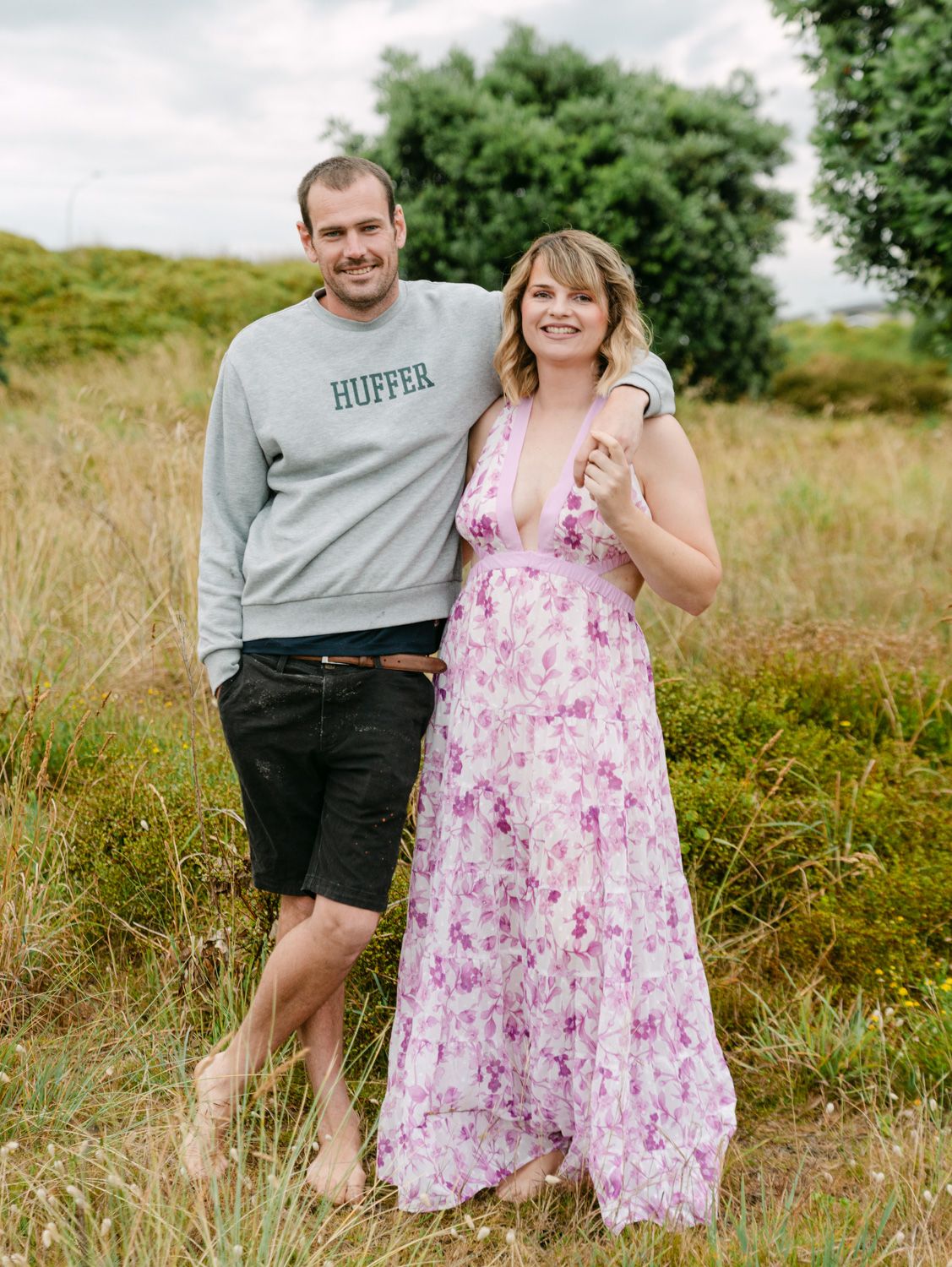 Couple posing outdoors in a grassy field, man in Huffer sweatshirt and shorts, woman in floral maxi dress.
