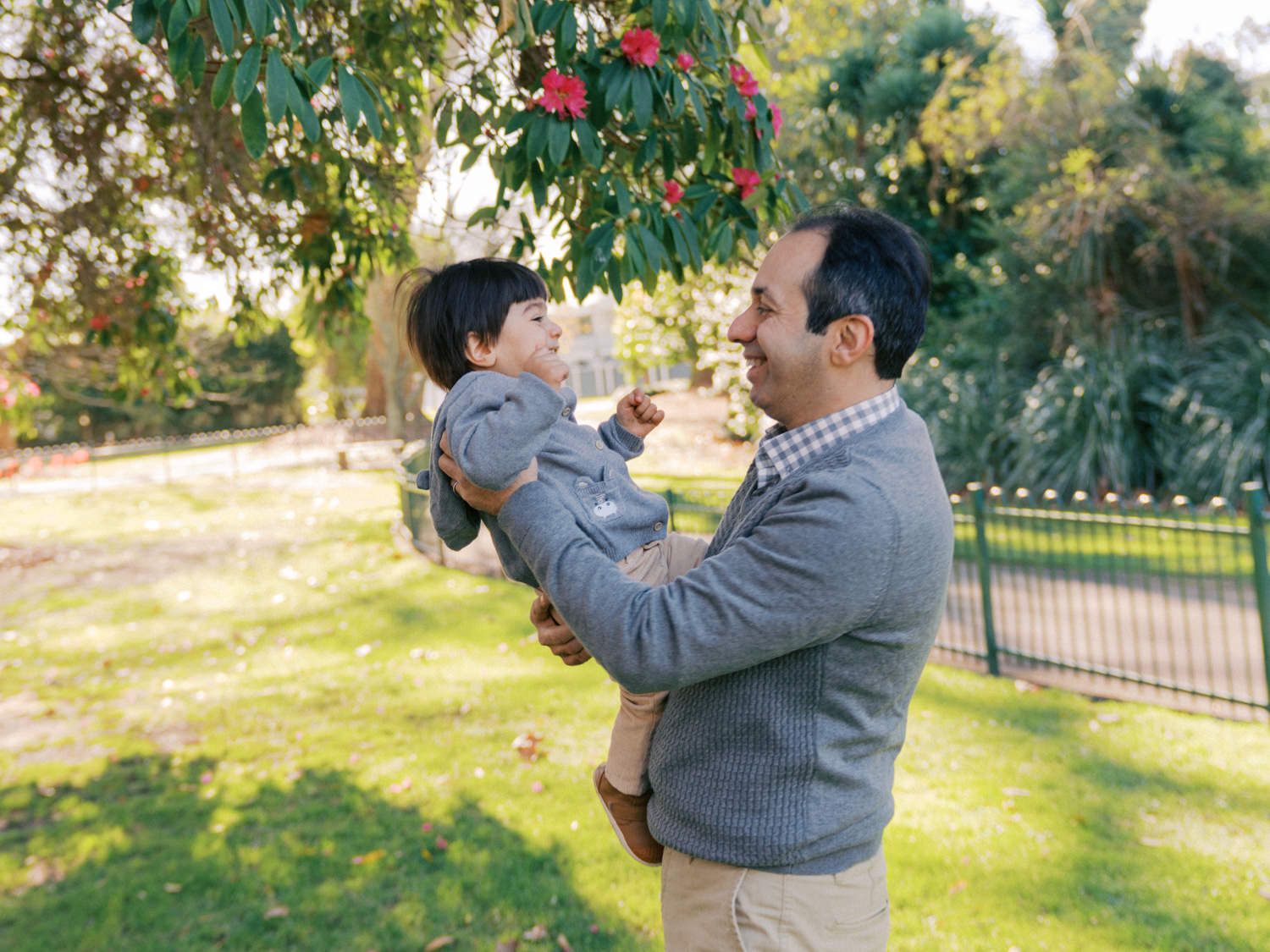 A father lovingly holds his toddler under a flowering tree in a lush green park.