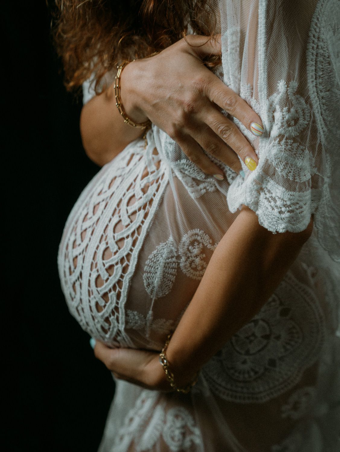 Pregnant woman in white lace maternity gown cradling baby bump, wearing gold bracelets, dark background.