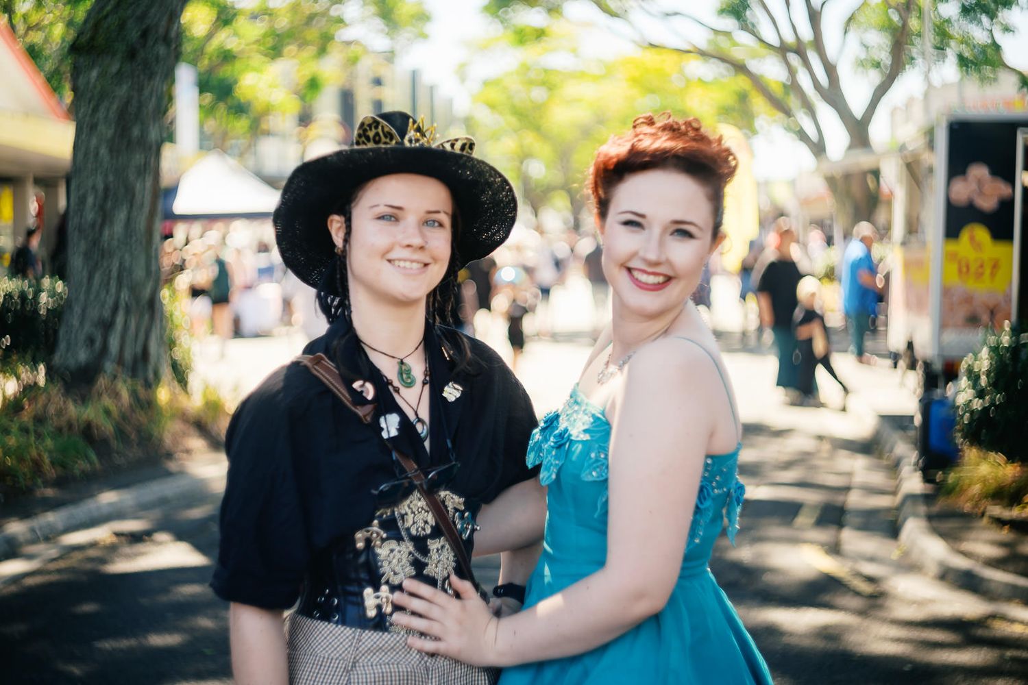 Two women in costume attire pose together outdoors at a festival, one in a steampunk hat and one in a blue gown.