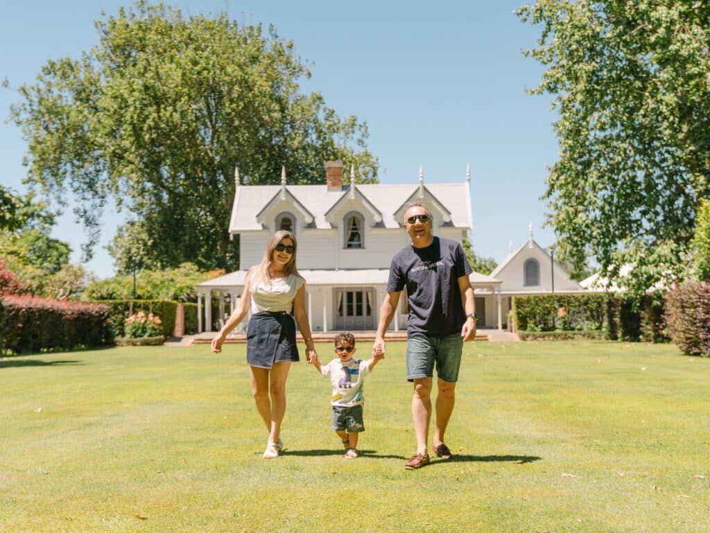Family of three holding hands walking on green lawn in front of a charming white country house on a sunny day.