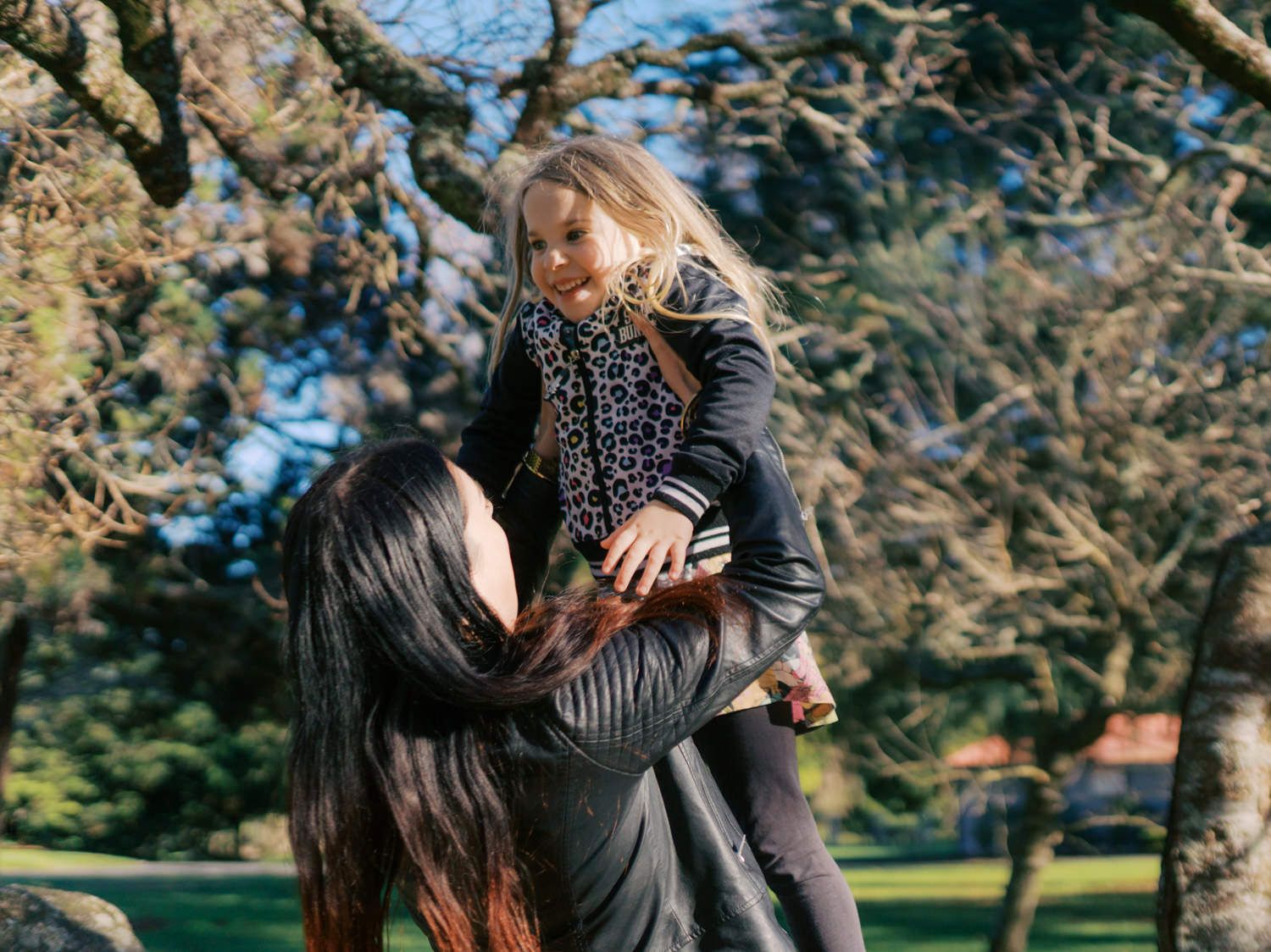 Woman lifts a laughing blonde girl into the air outdoors near a large tree in a park.