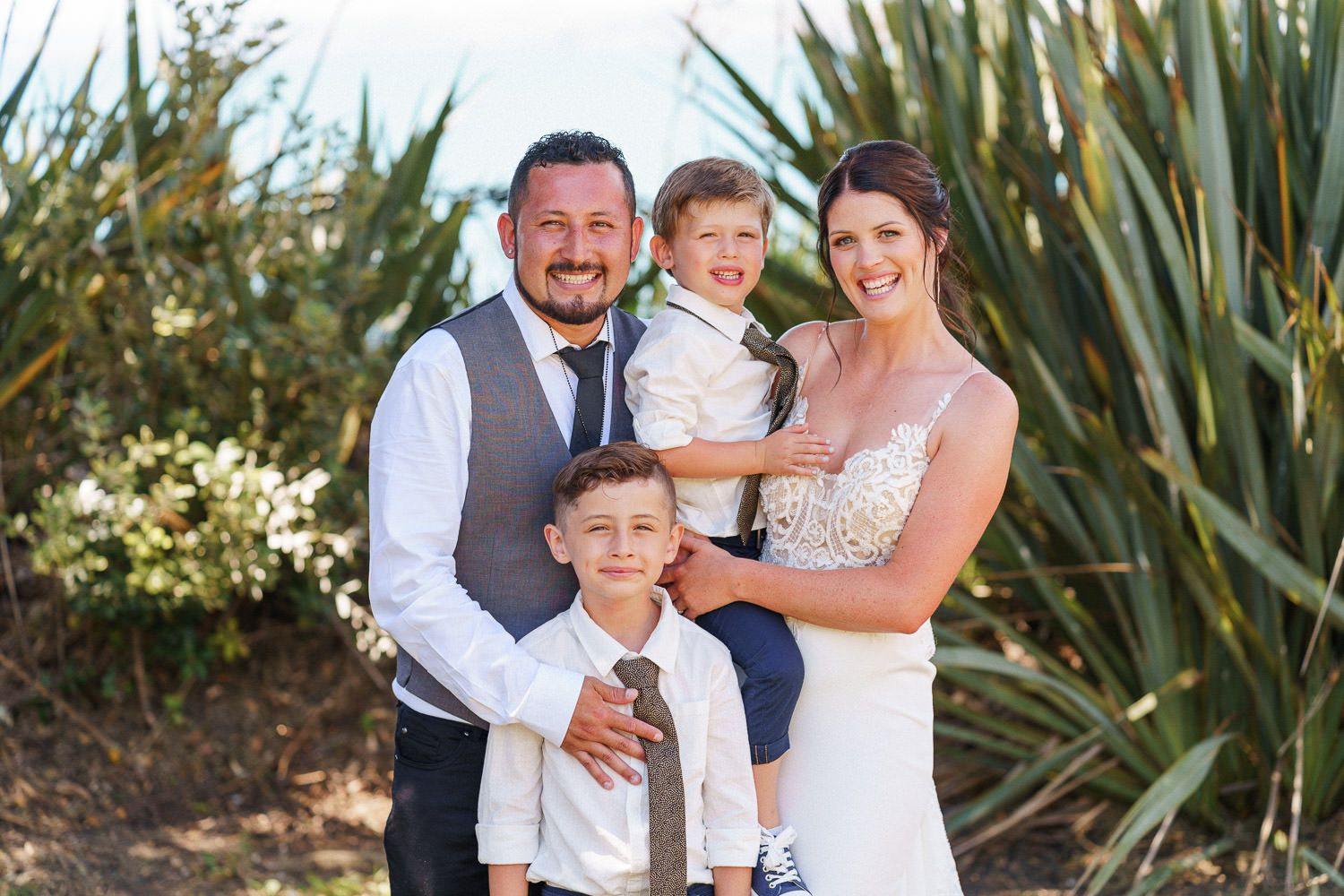 A happy family of four poses outdoors on a wedding day, with parents in formal attire and two young boys smiling.