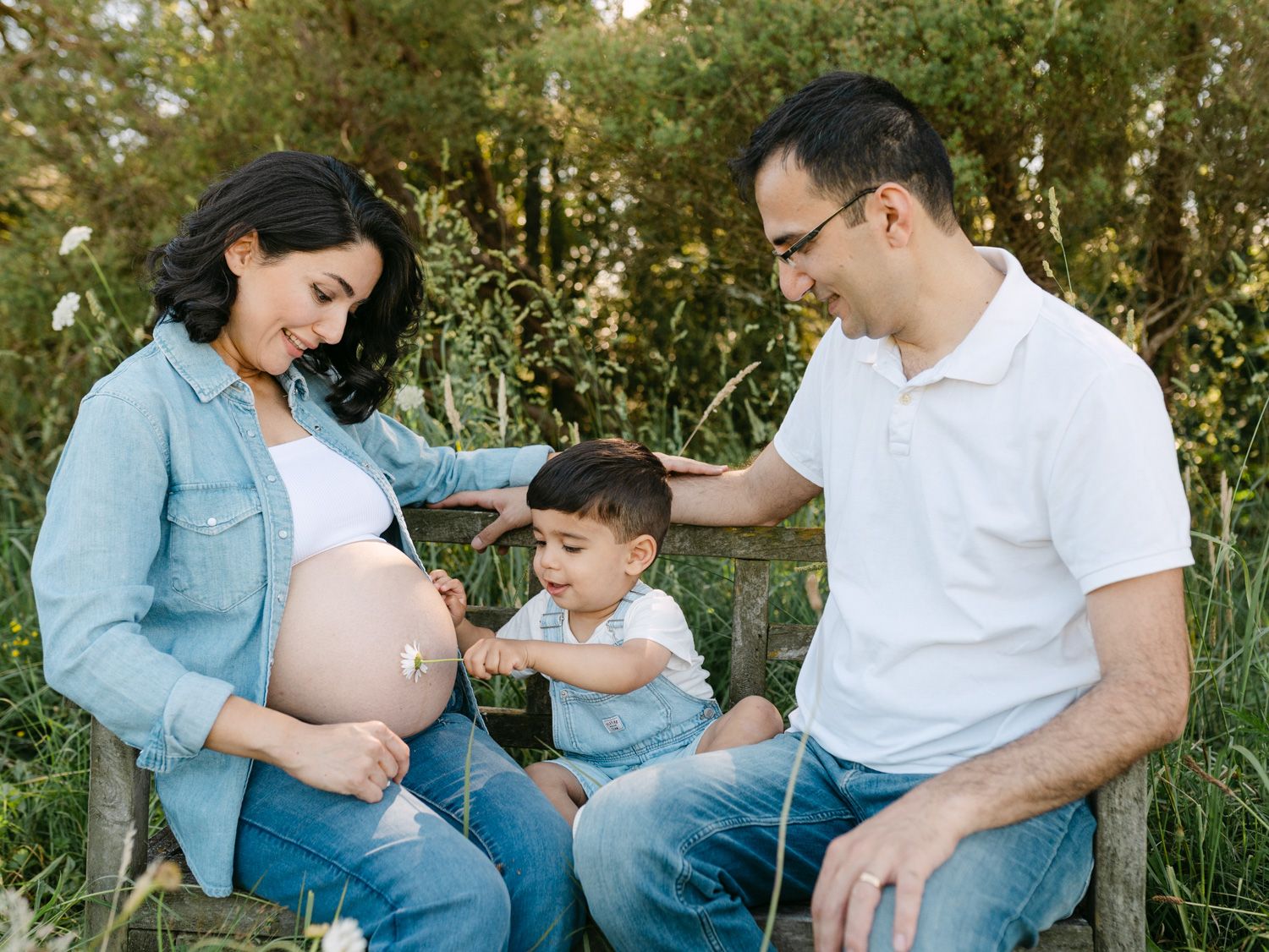 Pregnant woman sitting outdoors with husband and toddler son touching her baby bump during a family maternity photo session.