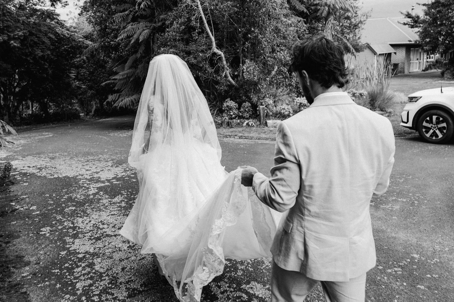 Black and white photo of a bride in a white gown and veil walking with a suited groom on a tree-lined path.