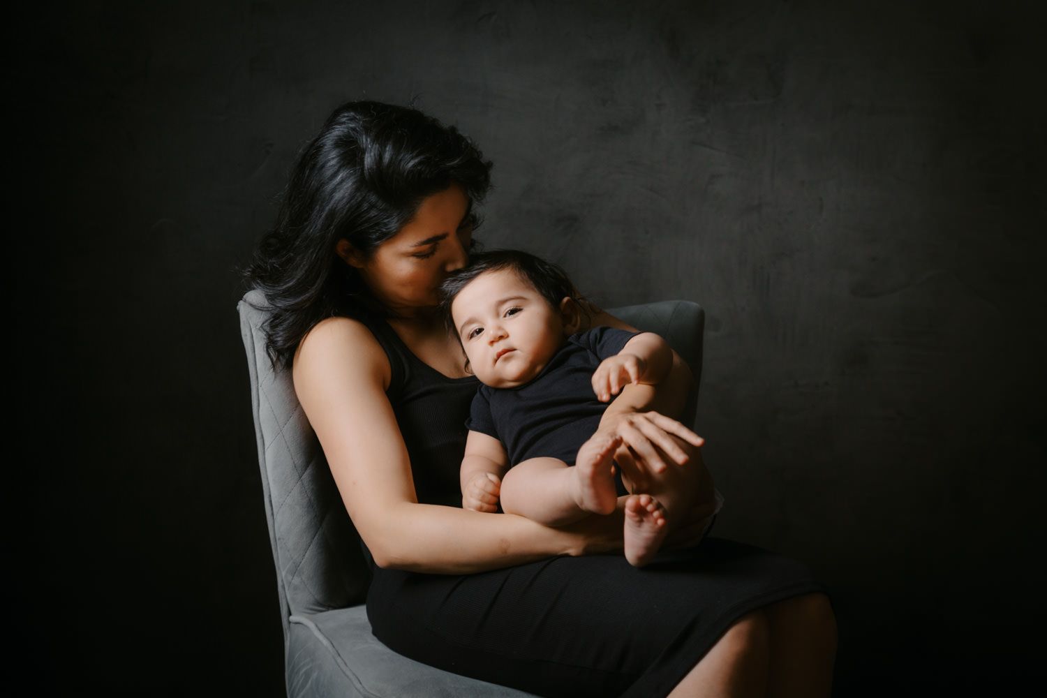 A mother in a black dress sits in a gray chair cradling her baby in a dark studio portrait.