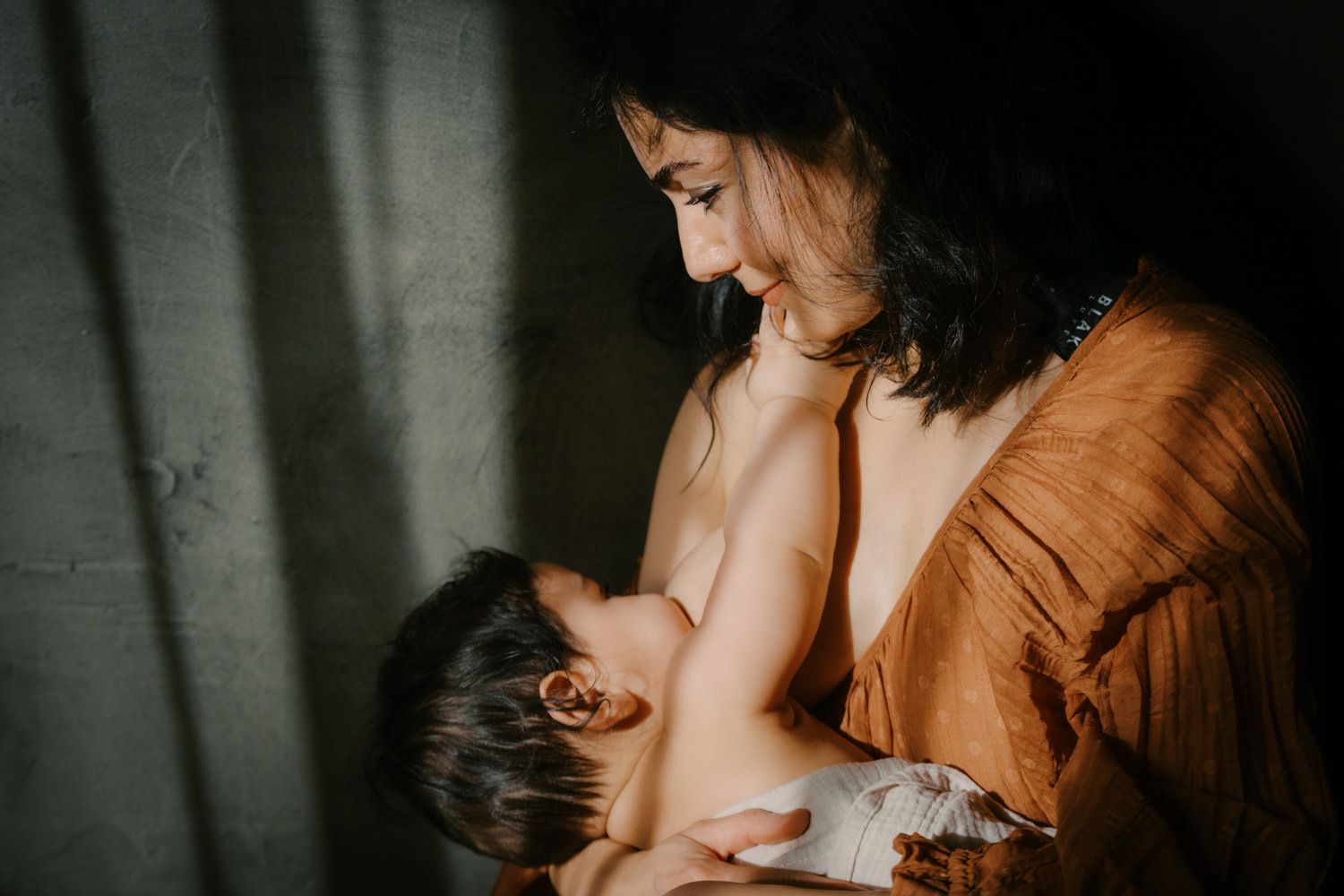 A mother tenderly breastfeeding her baby in warm natural light, wearing a rust-colored dress.